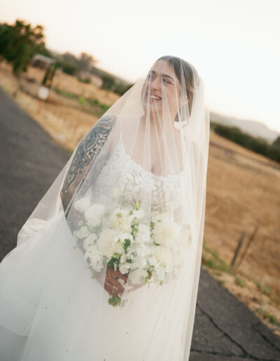 Bride in a white dress holding a bouquet of white flowers, standing on a rural road at sunset, with a veil covering her face and shoulders.