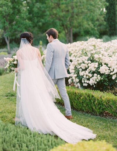 A bride and groom walk through a garden, with the bride in a white dress and veil and the groom in a light gray suit. They are surrounded by blooming white flowers and greenery.