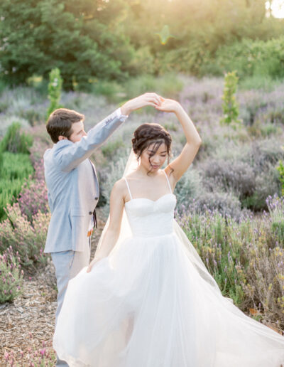 A couple dances in a garden with blooming flowers. The woman wears a white wedding dress, and the man is in a light gray suit. Sunlight filters through the trees.