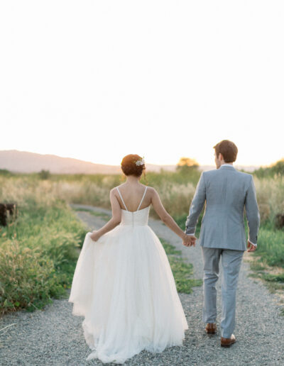 A couple holding hands walks down a gravel path during sunset. The woman wears a white dress, and the man wears a light gray suit.