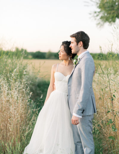 A couple in wedding attire stands in a grassy field, gazing into the distance under a pale sky.