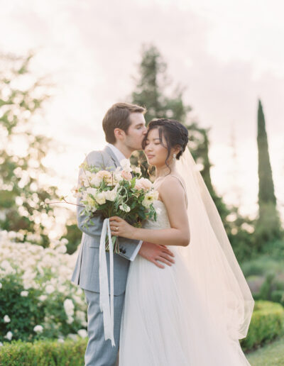 A couple in wedding attire embraces in a garden. The groom kisses the bride's forehead while she holds a bouquet. The setting is soft and romantic with trees in the background.