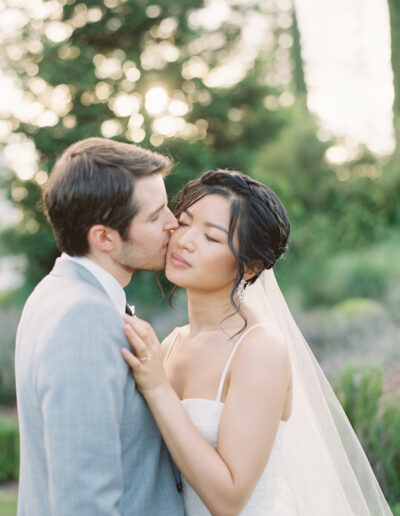 A bride and groom share an intimate moment outdoors, with the groom gently kissing the bride's cheek. They are dressed in wedding attire, surrounded by greenery and soft sunlight.