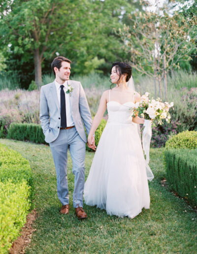 A bride and groom walk hand in hand down a garden path. The bride holds a bouquet, and both smile at each other.