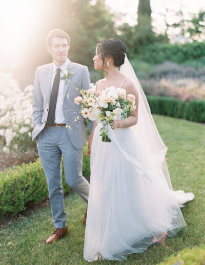 A bride and groom in a garden setting. The bride holds a bouquet and wears a flowing dress. The groom is dressed in a suit and tie, walking beside her.