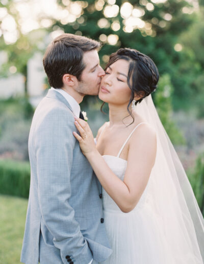 A groom in a light gray suit kisses the cheek of a bride in a white dress and veil, standing outdoors with greenery in the background.