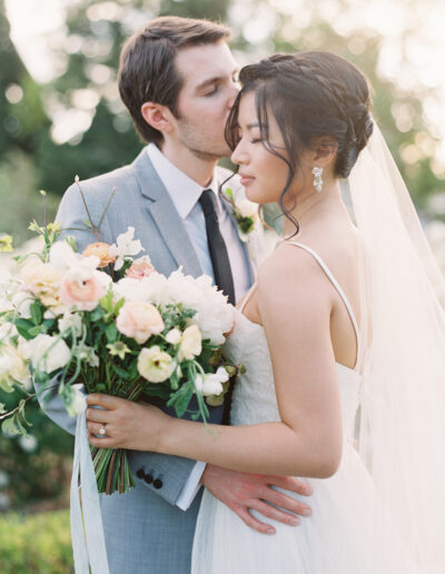 A groom in a light gray suit kisses the bride on her forehead. The bride holds a bouquet of white and blush flowers, wearing a white dress and veil. They're outdoors with greenery in the background.