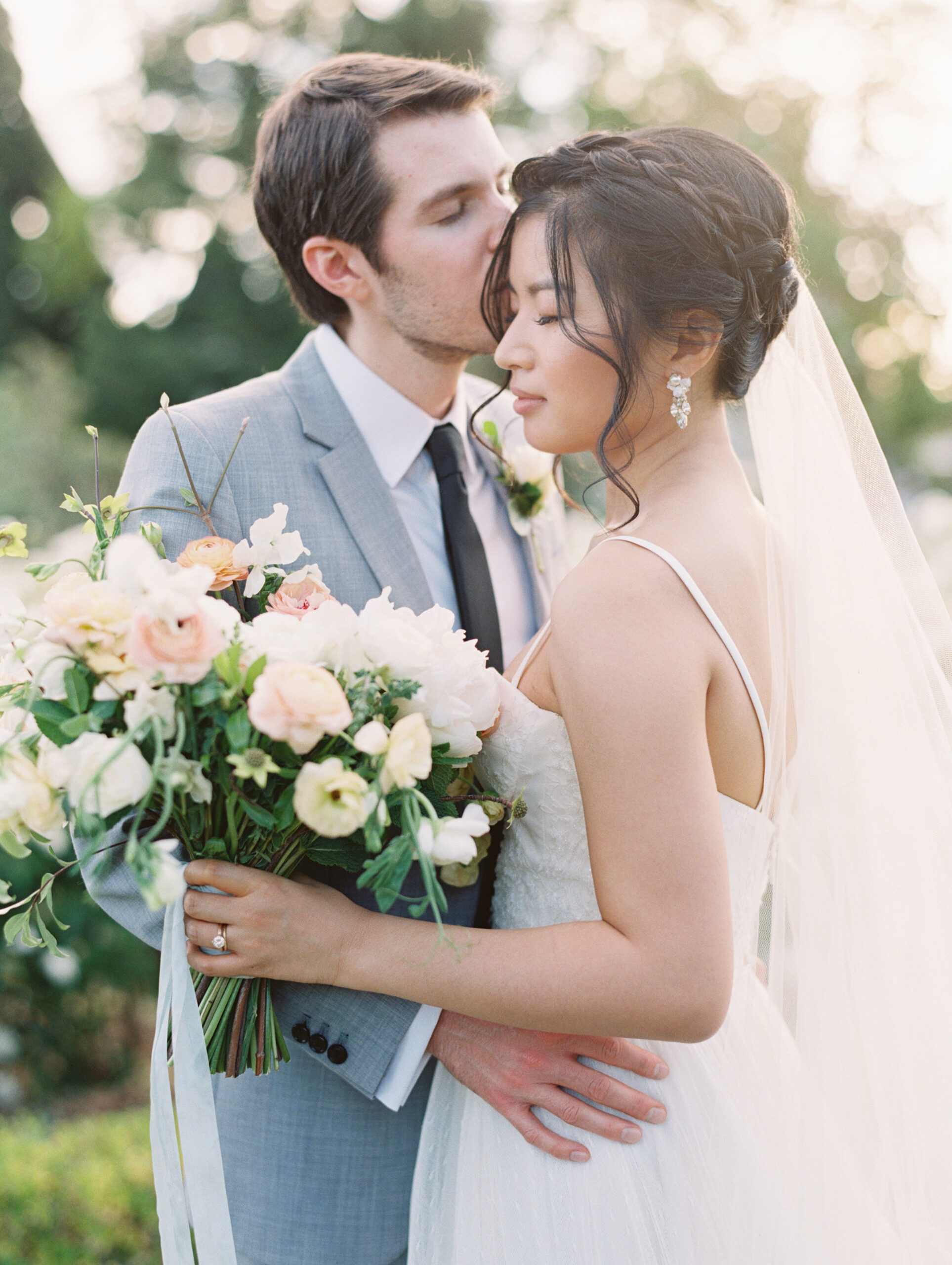 A groom in a gray suit kisses the forehead of a bride holding a bouquet of flowers. The bride wears a white dress and veil.