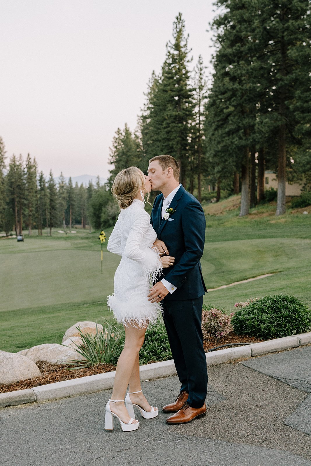 A couple in formal attire kisses outdoors near a golf course, surrounded by trees.