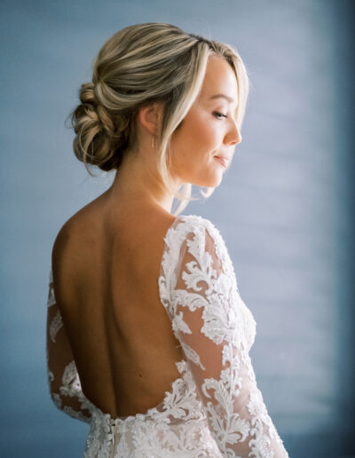 A woman in an intricately patterned white lace dress with an open back and her hair styled in an updo stands against a soft blue background, looking to the side.