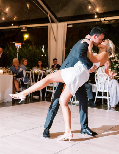 A couple in formal attire perform a dance at a wedding reception, with the woman lifted and dipped by the man. Guests are seated at tables in the background under a tent with string lights.