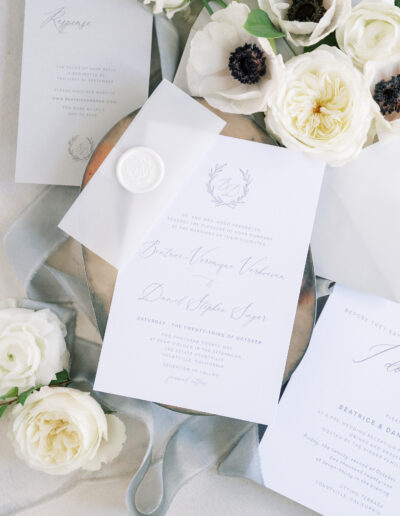 Elegant wedding invitation suite with floral designs, surrounded by white roses and greenery on a soft textured background.