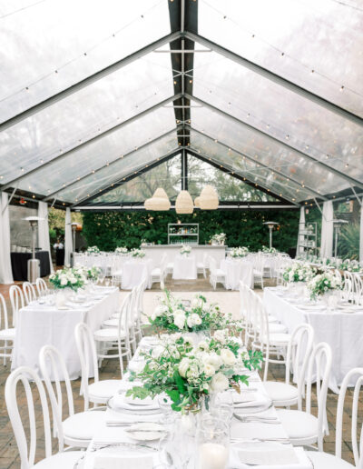 Elegant outdoor wedding reception setup under a clear tent, featuring white tables and chairs with green and white floral centerpieces. Hanging pendant lights create a warm atmosphere.