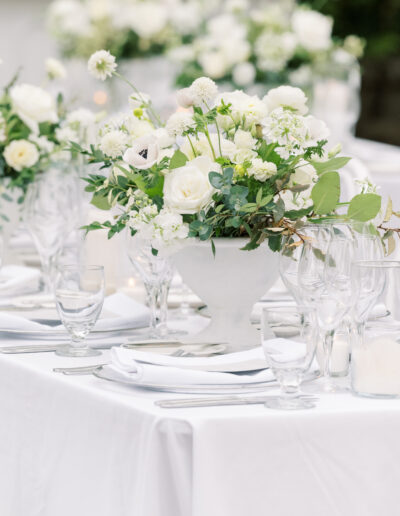 A white tablecloth-covered table is elegantly set with white floral centerpieces, glassware, and neatly folded napkins.