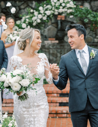 Bride and groom walk down the aisle holding hands, surrounded by guests. The bride holds a bouquet, and guests are throwing flower petals.