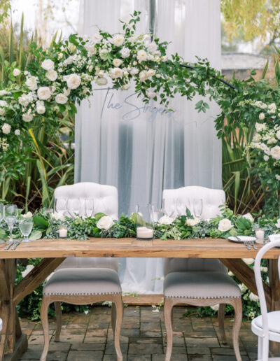 Elegant wedding table with two white chairs, adorned with white flowers and greenery, set under an arch of similar floral arrangement.