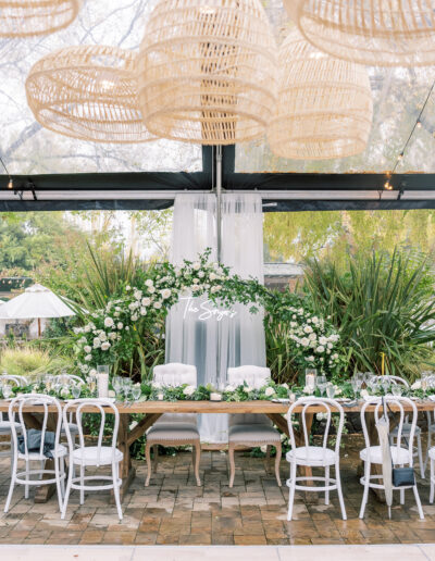 Elegant wedding table setting with white chairs, greenery, and floral centerpiece under hanging woven lamps. A sheer curtain backdrop with "The Sages" written on it.