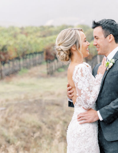 A bride and groom in wedding attire embrace joyfully in an outdoor vineyard setting on a misty day.