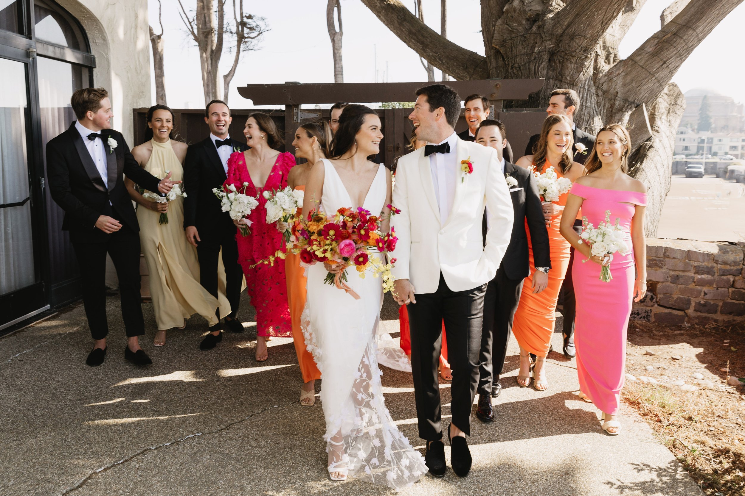 A bride and groom in wedding attire walk outdoors with a group of bridesmaids and groomsmen in colorful dresses and suits, holding flowers.