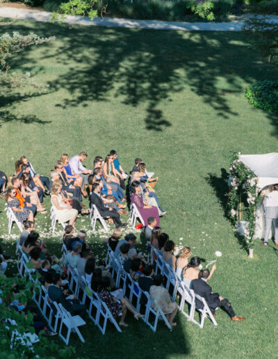 Outdoor wedding ceremony with guests seated on chairs, facing a couple under a floral canopy. The setting is grassy with trees providing shade.