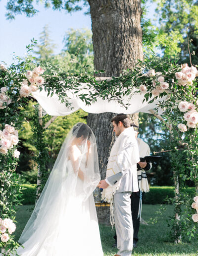 A bride and groom stand under a floral wedding arch outdoors, with a large tree in the background. The groom holds a cloth, and both are dressed in formal attire.