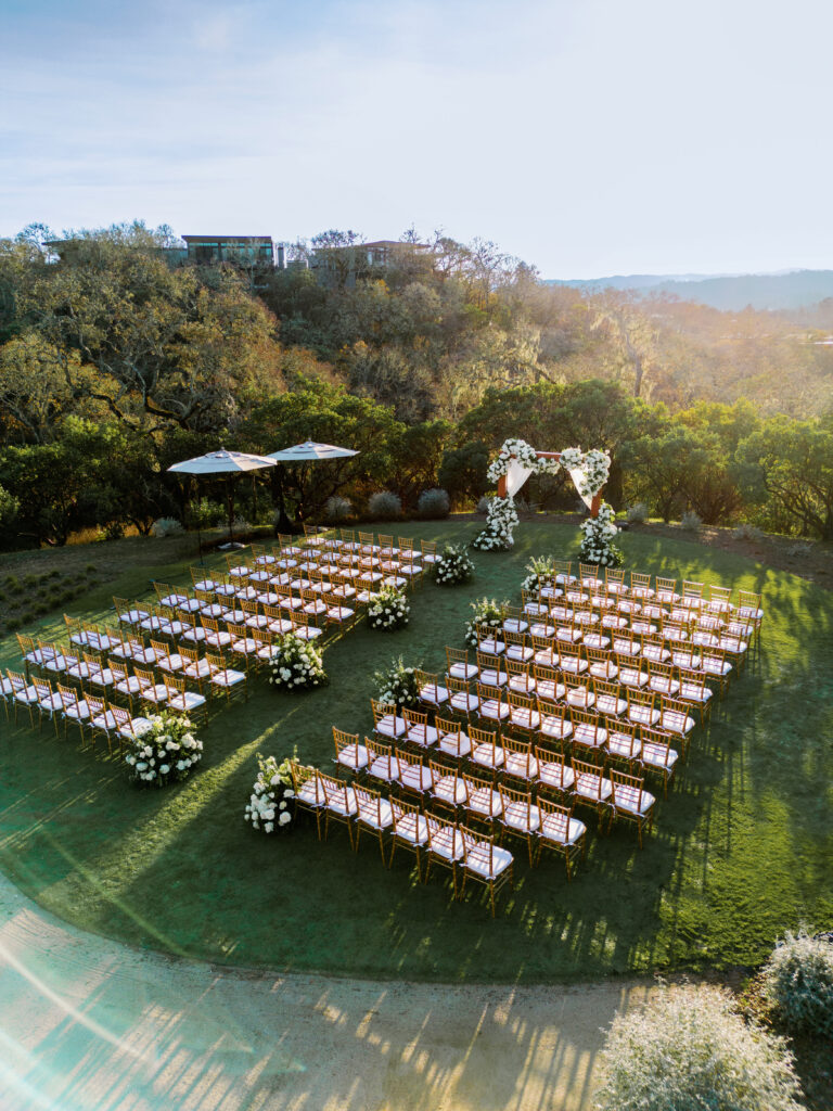 Outdoor wedding setup with rows of wooden chairs, floral arrangements, and a heart-shaped arch. Trees and hills are in the background.