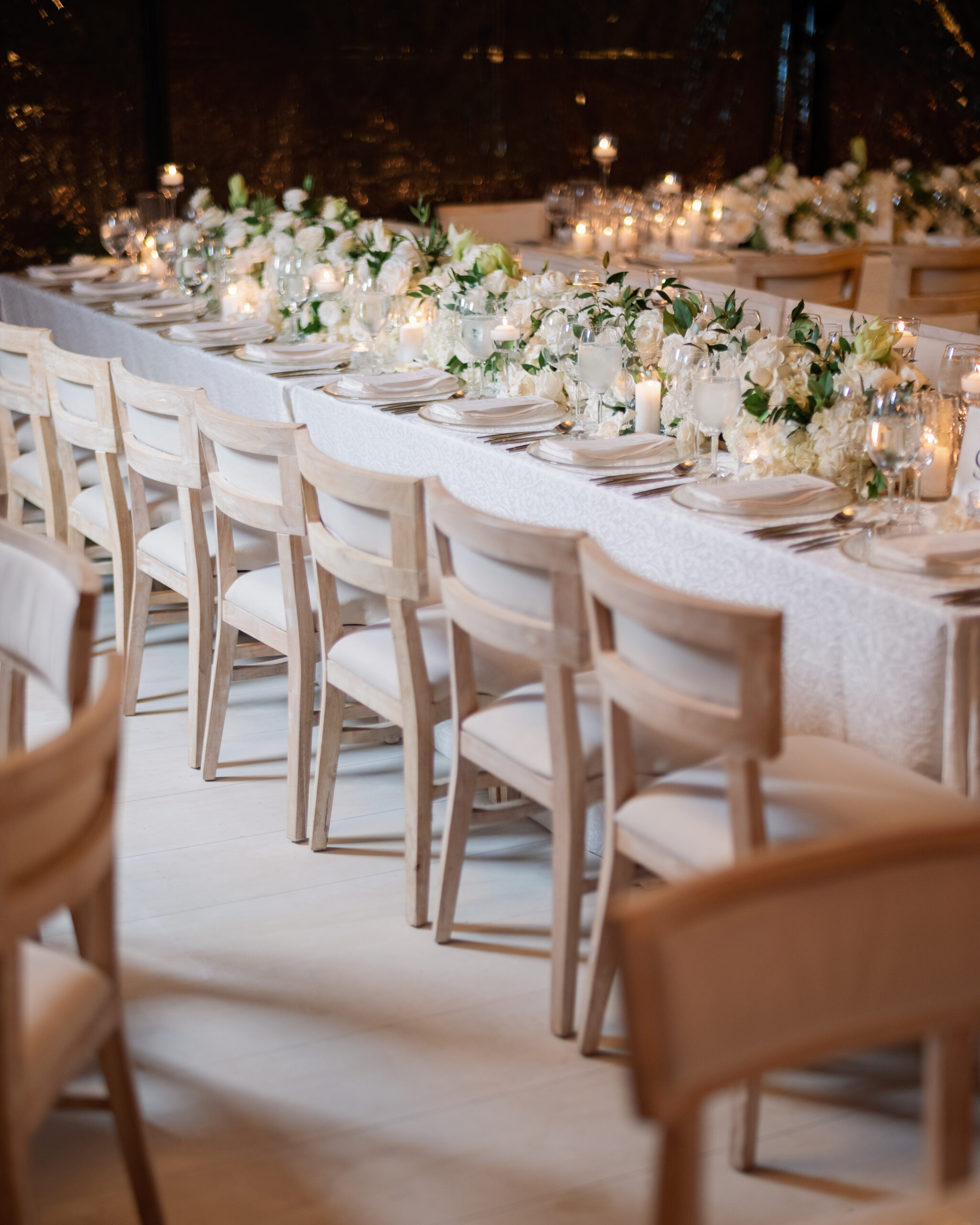 A long dining table set with white tablecloth, floral centerpieces, candles, and neatly arranged wooden chairs in an elegant setting.