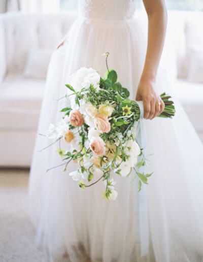 A bride in a white dress holds a bouquet of white and pink flowers with greenery, standing in a softly lit room.