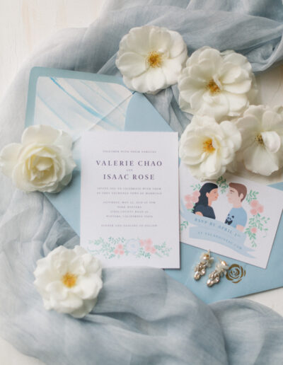 Flat lay of wedding invitations and RSVP card on a light blue fabric, surrounded by white flowers and pearl earrings.