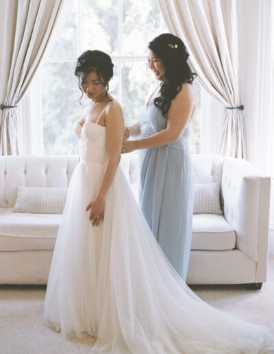 A bride in a white gown stands while another woman in a light blue dress adjusts her dress in front of a sofa by a window.