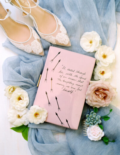 Flat lay of a pink book with a quote, surrounded by white and blush flowers, a light blue cloth, and white lace high-heeled shoes.