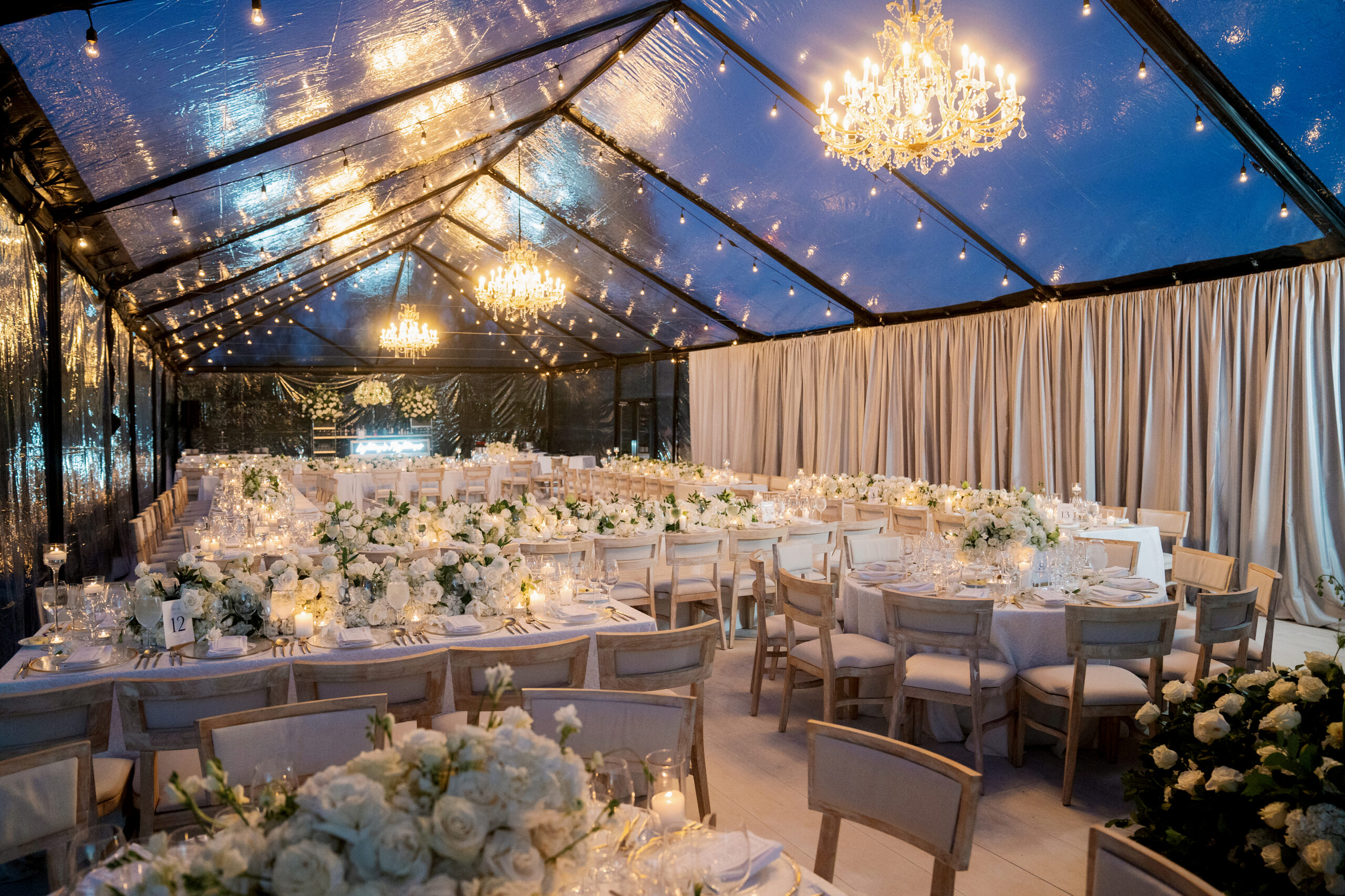 A beautifully decorated Montage Healdsburg wedding reception features round tables, white chairs, and floral centerpieces. Under a transparent tent adorned with string lights and chandeliers, this enchanting indoor setting captures the essence of a dream celebration.