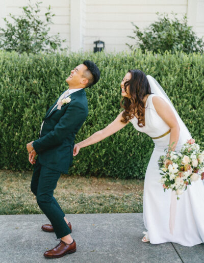 A bride playfully tugs at the groom's jacket, both laughing, in front of greenery. She holds a bouquet of flowers.