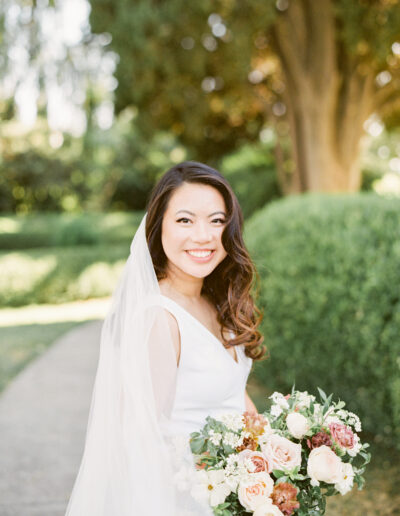 A bride in a white dress smiles while holding a bouquet of flowers. She is standing outdoors with greenery in the background.