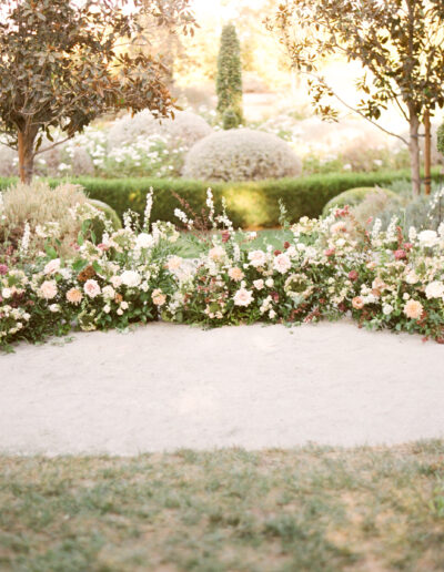 A circular arrangement of flowers and greenery in a garden setting with manicured bushes and trees, bathed in soft sunlight.