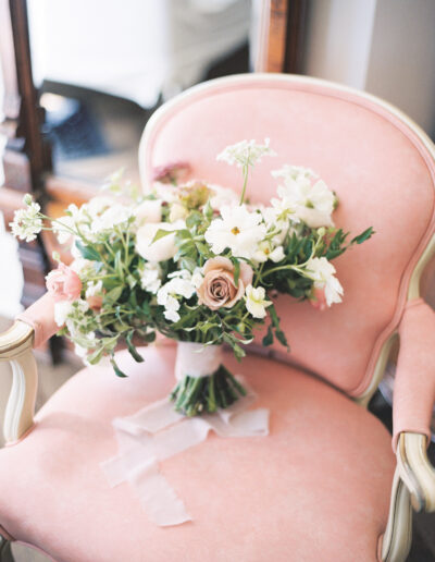 A bouquet of flowers with roses and greenery rests on a pale pink upholstered chair.