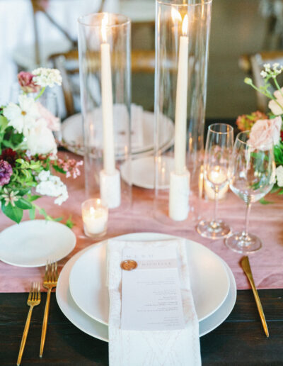 Elegant table setting with white plates, a menu, gold cutlery, and glasses. Tall candles in glass holders and floral arrangements decorate the table on a pink runner.