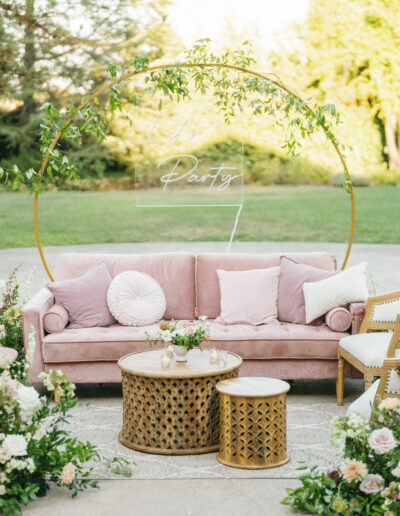 Outdoor seating area with a pink sofa, chairs, and floral arrangements, featuring a circular frame with greenery.