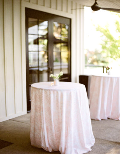 Round table with a white cloth and small flower vase on a patio. Another similar table is in the background near a glass door.