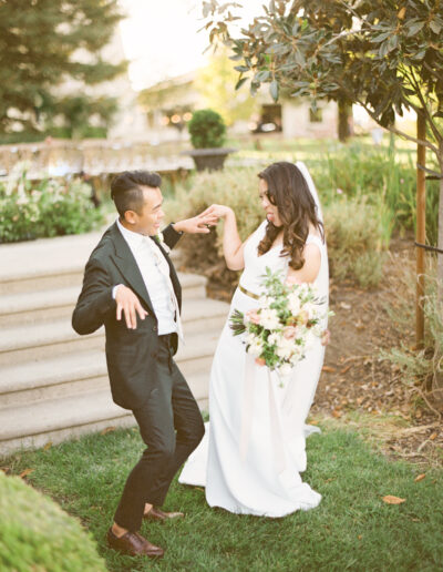 A groom and bride in a playful pose outdoors, with the bride holding a bouquet and both dressed in formal attire.