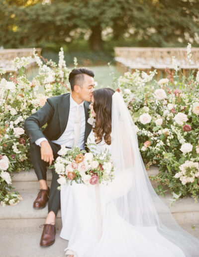 A bride and groom sit on steps surrounded by flowers. The groom is in a suit, and the bride wears a veil, holding a bouquet. They lean closely together.