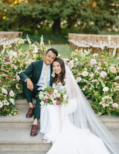 A couple in wedding attire sits on steps surrounded by lush floral arrangements and greenery.