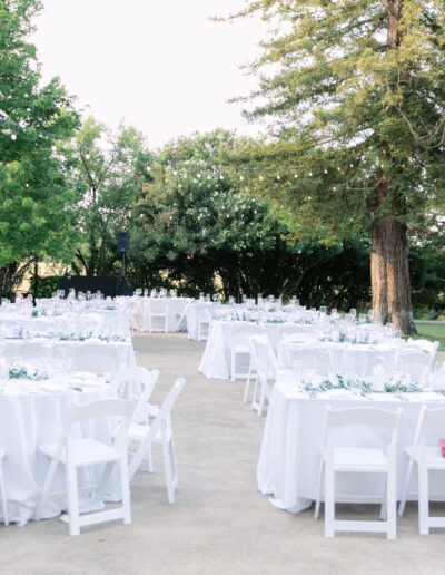 Outdoor event setup with round tables covered in white tablecloths, surrounded by white chairs, in a garden setting with trees.