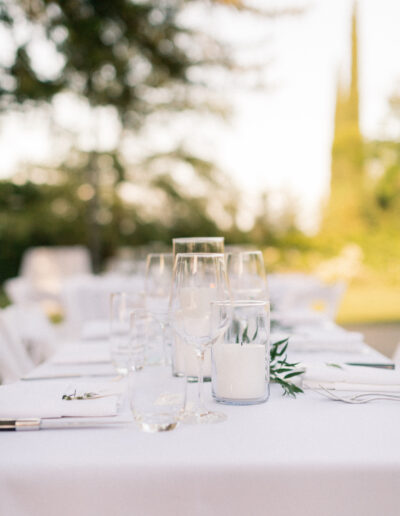 A close-up of a white table set elegantly outdoors with glasses and candles, surrounded by greenery in the background.