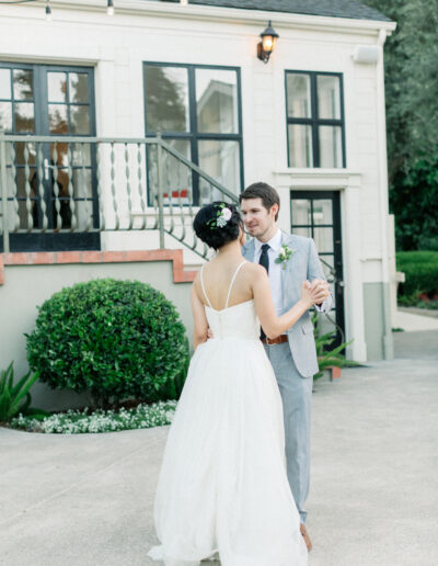 A couple dances outside, the woman in a white dress and the man in a gray suit, in front of a house with string lights.