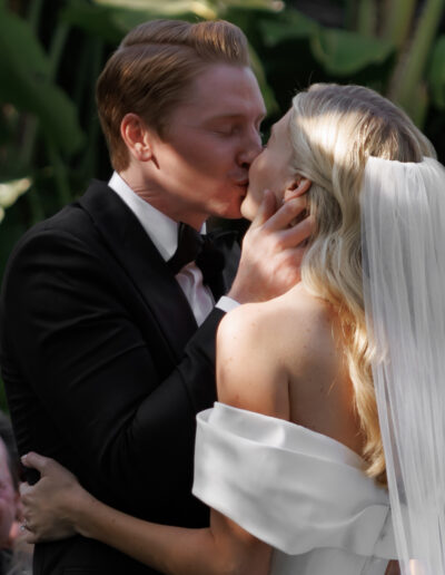 A couple in wedding attire share a kiss outdoors; the groom wears a black suit, and the bride wears a white dress with a veil.