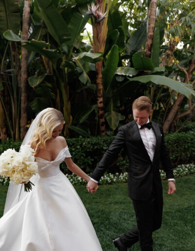 A bride in a white dress holds a bouquet of white flowers while walking hand in hand with a groom in a black suit, in a garden setting.