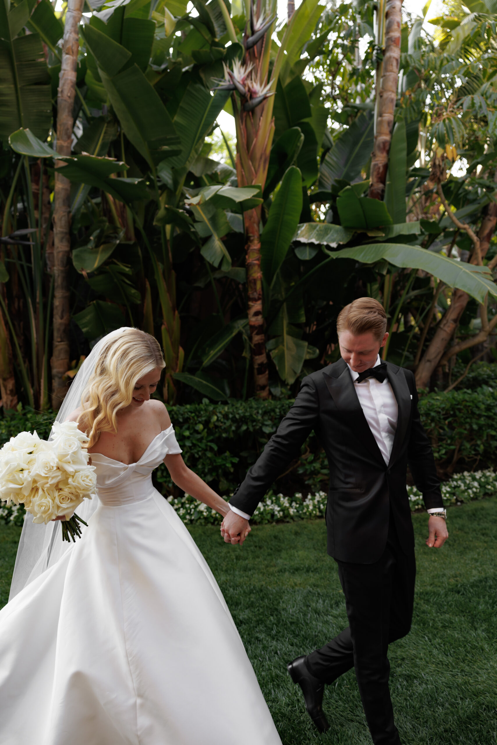 A bride in a white dress holds a bouquet of white flowers while walking hand in hand with a groom in a black suit, in a garden setting.