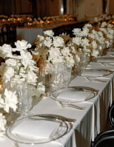 A table set for an event with white tablecloth, glassware, plates, and white floral arrangements in vases.