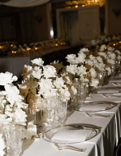 Elegant table setting with white roses, crystal glassware, and white plates on a long draped table. Candles are interspersed among the flowers, creating a formal dining ambiance.